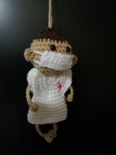 Gehäkelte Amigurumi-Puppe mit Mundschutz und kleinem roten Kreuz‑Symbol auf weißem Oberteil, hängend vor dunklem Hintergrund.