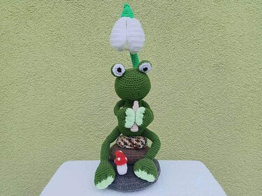 Gehäkelter grüner Amigurumi-Frosch mit großen Augen, weißem Schneeglöckchen auf dem Kopf, hellgrünem Schmetterling in den Händen und kleinem roten Pilz auf grauem Sockel.