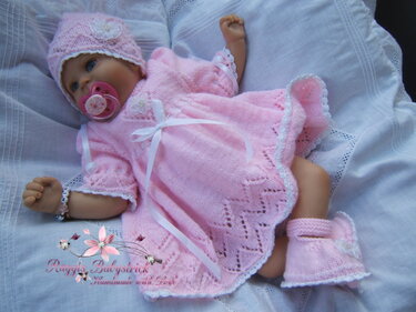Babypuppe in rosa Strickkleid mit Lochmuster, passender Mütze und Booties, mit weißer Spitze und Satinschleife.