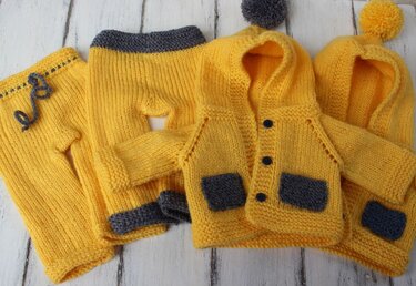 Two yellow knitted hooded cardigans with grey pockets and buttons, two pairs of yellow knitted pants with grey trim, and two yellow hats with pompoms, laid on a white wooden surface.