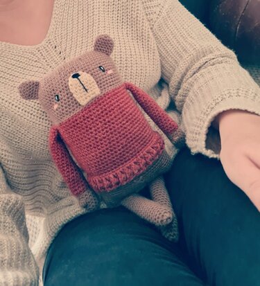 Gehäkelter brauner Amigurumi-Teddybär mit rotem Pullover auf dem Schoß.