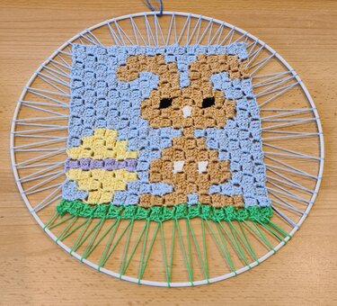 Gehäkeltes Quadrat mit Hasen‑ und Ostereimotiv, in einem runden Metallring gespannt.