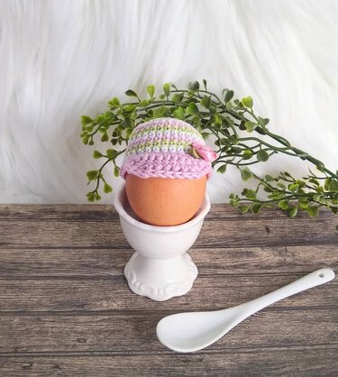 Braunes Ei im weißen Eierbecher mit gehäkeltem Eierwärmer als Schirmmütze in Pastellfarben mit kleiner Blüte