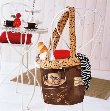 Geräumige braune Einkaufstasche aus Stoff mit Kaffeebohnenmuster, Zebra-Tassenmotiv und einem Baguette, hängt an einem weißen Stuhl.
