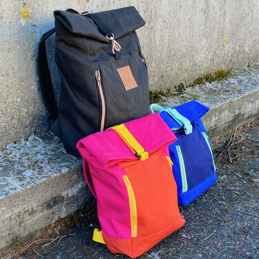 Drei Rolltop-Rucksäcke in Schwarz, Pink/Orange und Blau nebeneinander an einer Betonmauer