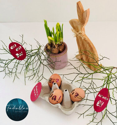 Osterdekoration mit Holzhase, Narzissen, beschrifteten Eiern und roten ausgeschnittenen Plott-Anhängern an Zweigen