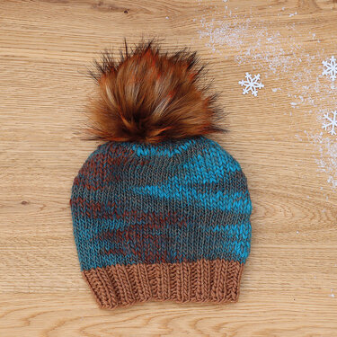 Melierte Strickmütze in Blau, Grau und Braun mit braunem Kunstfellbommel auf Holztisch neben Deko-Schneeflocken