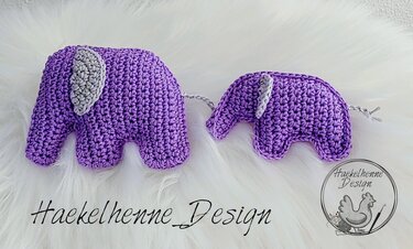 Zwei handgehäkelte lila Amigurumi-Elefanten mit grauen Ohren und Schwänzchen auf weißem, flauschigem Hintergrund