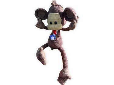 Gehäkelter brauner Amigurumi-Affe mit beigem Gesicht und blau-rotem Anhänger am Hals vor weißem Hintergrund.