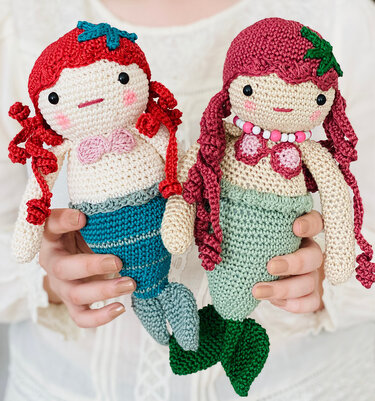 Zwei gehäkelte Amigurumi-Meerjungfrauen in Rot und Magenta, gehalten von einer Person.