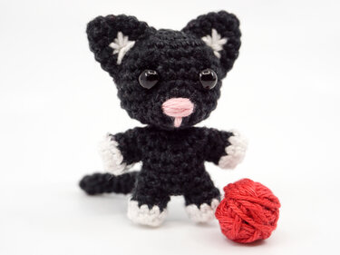 Small black crocheted amigurumi cat with white paws and pink nose sitting beside a red yarn ball on a white background