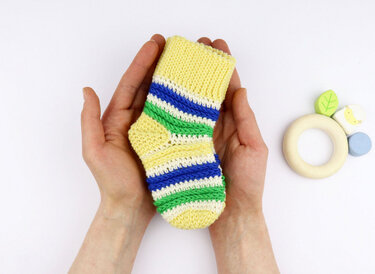 Gehäkelte Babysocke in Gelb mit blauen, grünen und weißen Streifen, gehalten in zwei Händen, neben einem Holzspielzeug auf weißem Hintergrund.