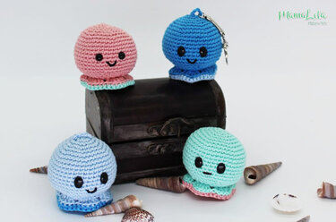 Vier gehäkelte Oktopus‑Amigurumis in Pastellfarben, einer mit Schlüsselring, neben einer kleinen Schatztruhe und Muscheln auf weißem Hintergrund.