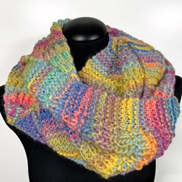 Mehrfarbiger gestrickter Loop-Schal in Pastelltönen (Gelb, Rosa, Blau, Grün) auf schwarzer Schaufensterpuppe vor hellem Hintergrund.