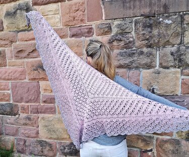 Gehäkeltes zartrosa Dreieckstuch mit Lochmuster und dekorativer Borte, getragen vor einer Steinwand.