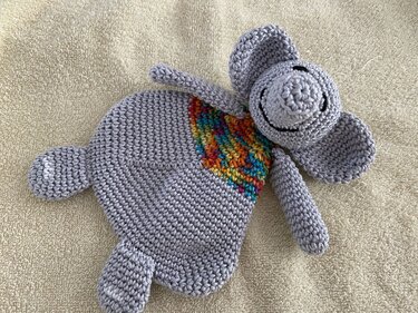 Gehäkeltes graues Elefanten‑Schmusetuch mit buntem regenbogenfarbenem Einsatz auf hellem Textil liegend