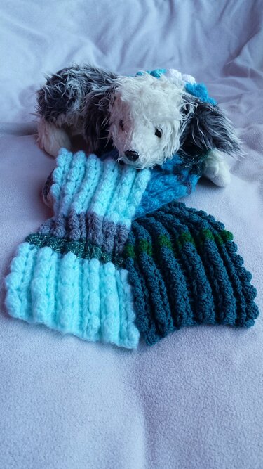 Gehäkelter Kinderschal mit horizontalen Streifen in Hellblau, Dunkelblau, Grau und Grün auf einer hellen Decke neben einem Plüschtier