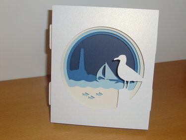 Weiße Klappkarte mit kreisförmigem Ausschnitt und mehrlagiger maritimer Papier-Szene mit Leuchtturm, Segelboot, Möwe und Fischen
