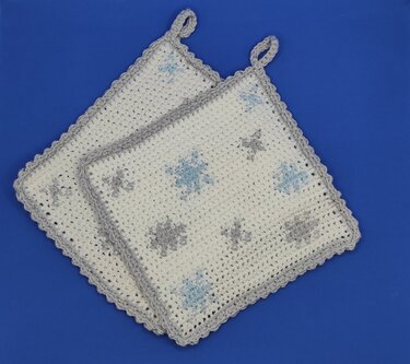 Zwei weiße gehäkelte Topflappen mit hellblauen und grauen Sternen, gewelltem Rand und Aufhängeschlaufen