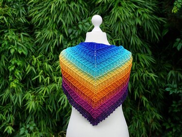 Gehäkeltes Dreieckstuch in Regenbogenfarben auf weißer Schneiderpuppe vor grünen Blättern.