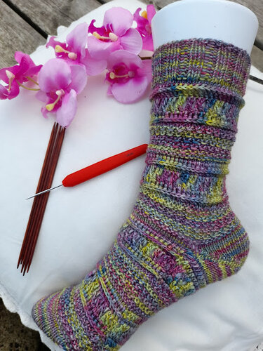 Bunte, handgemachte Socke auf weißem Sockenspanner, daneben Nadelspiel, rote Häkelnadel und rosa Orchideen.