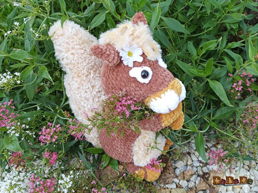 Flauschiges Amigurumi-Eichhörnchen in Braun und Beige mit buschigem hellem Schwanz und weißer Blume am Kopf zwischen grünen Blättern