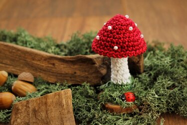 Roter, gehäkelter Fliegenpilz mit weißen Perlenpunkten auf Moos, neben Eicheln und Marienkäfer.