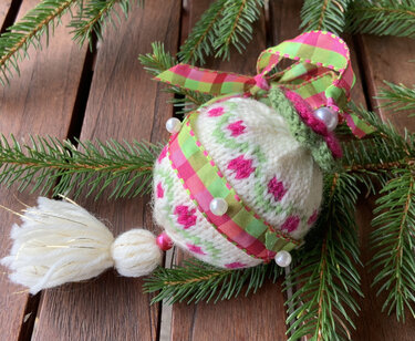 Gestrickte Weihnachtskugel in Weiß, Pink und Grün mit kariertem Band, Perlen und Quaste auf Holztisch und Tannengrün