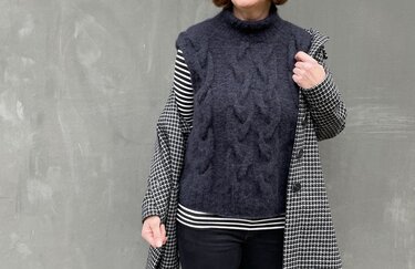 Ärmellose dunkelgraue Strickweste mit Zopfmuster und Stehkragen, getragen über gestreiftem Langarmshirt; kariertes Mantelteil über der Schulter.