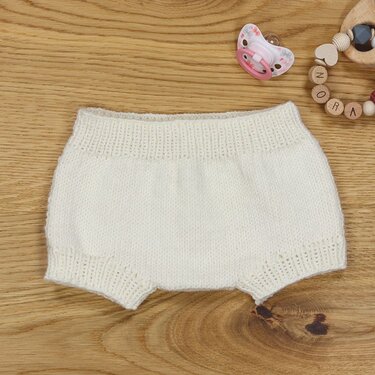 Cremefarbene gestrickte Babyhose auf Holzuntergrund neben Schnuller und Greifling