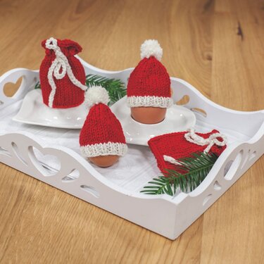 Rote gestrickte Eierwärmer als Weihnachtsmützen mit weißen Bommeln und zwei rote Geschenktaschen auf einem weißen Tablett mit Tannengrün.