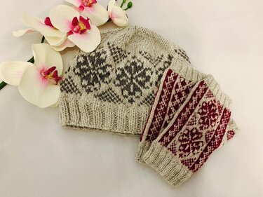 Beige gestrickte Mütze mit grauem Ornamentmuster und beige-rote Stulpen auf hellem Hintergrund neben Orchideen
