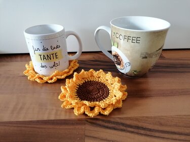 Zwei gehäkelte Untersetzer in Sonnenblumenoptik (braune Mitte, gelbe Rüschen) neben zwei Kaffeetassen auf Holz