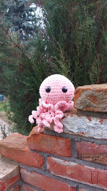 Gehäkelte rosa Amigurumi-Qualle mit großen schwarzen Augen auf einer Ziegelmauer vor grünen Büschen.
