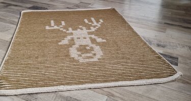 Beige gestrickte Babydecke mit weißem Rentierkopf und heller Kante auf Holzfußboden