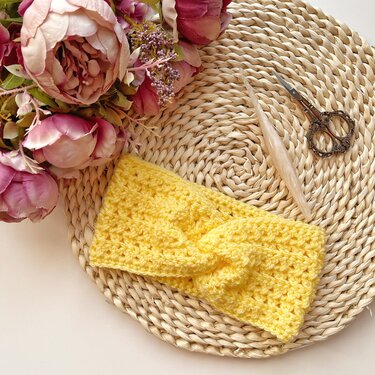 Yellow crocheted headband with twisted knot on woven mat beside pink flowers, a crochet hook and scissors.