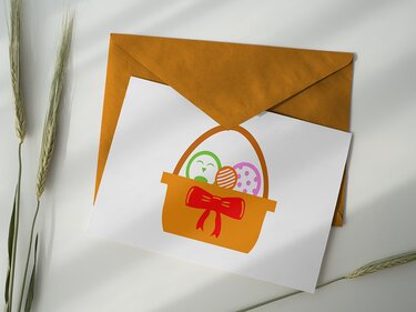 Weiße Karte in braunem Umschlag mit orangefarbenem Osterkorb, roter Schleife und drei bunten Ostereiern