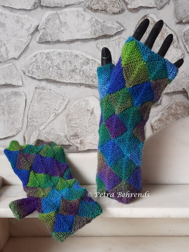 Bunte gestrickte fingerlose Damen-Armstulpen im Entrelac-Rautenmuster auf schwarzer Mannequin-Hand vor hellem Steinmauer-Hintergrund