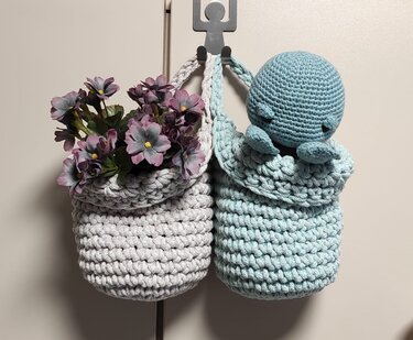 Zwei gehäkelte Hängekörbe, grau mit lila Kunstblumen und türkis mit gehäkeltem Oktopus, an einem Haken hängend.