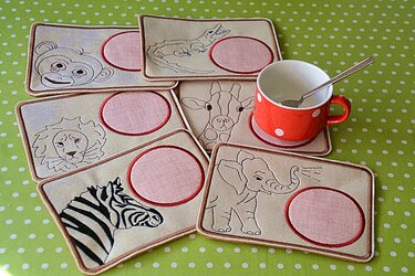 Sechs bestickte rechteckige Tassenteppiche mit skizzenhaften Tiermotiven auf grün gepunktetem Stoff; rosa Tassenflächen und eine rote Tasse mit Löffel auf einer Matte