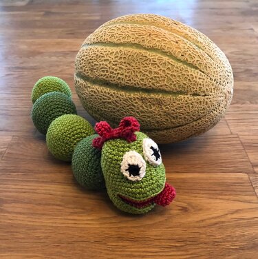 Handgehäkelte grüne Raupe mit roten Details und großen Augen neben einer Melone auf Holz