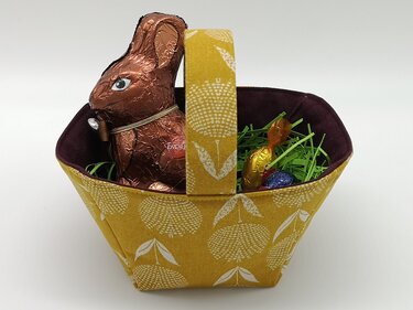 Gelber genähter Stoffkorb mit Henkel und weißem Blumenmuster, dunkler Innenfütterung, gefüllt mit einem Schokoladenosterhasen, grünem Ostergras und Süßigkeiten.