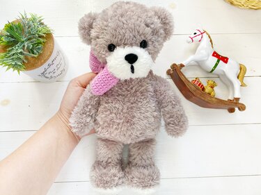 Hand holding a gray fluffy amigurumi teddy bear wearing a pink crocheted scarf on a white wooden surface with a succulent pot and a rocking horse nearby