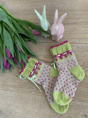Gestrickte Kindersocken in Beige mit rosa Punkten, hellgrüner Spitze und Ferse sowie eingezogenem Band, arrangiert mit Tulpen und Deko-Hasen.