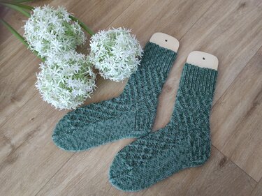 Dunkelgrüne handgestrickte Socken mit Rautenmuster auf hölzernen Sockenformern neben weißen Zierblumen.