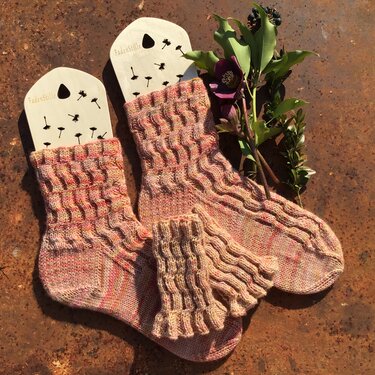 Handgestrickte rosa-melierte Socken und passende Handstulpe mit Wellenmuster auf Sockenspannern neben grünen Blättern