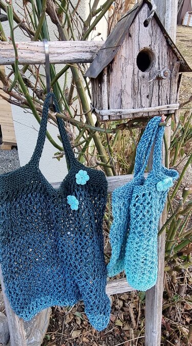 Zwei gestrickte Netztaschen in Blau und Türkis mit Blumenapplikationen, hängend an einer Holzlatte neben einem Vogelhaus