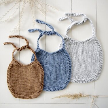 Set aus drei gestrickten Babylätzchen in Braun, Hellblau und Hellgrau auf wei
ßem Holz.