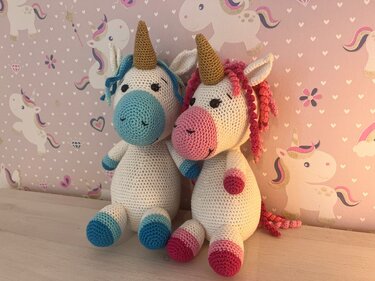 Zwei gehäkelte Einhorn-Amigurumi in Blau und Rosa mit goldenen Hörnern vor Einhorn-Tapete