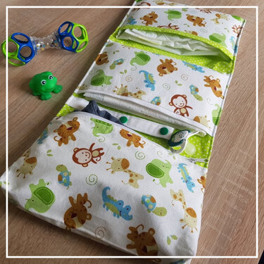 Gefaltete Windeltasche aus Stoff mit buntem Tiermuster, mehreren Fächern, grünen Druckknöpfen und Schnullerband auf Holzuntergrund